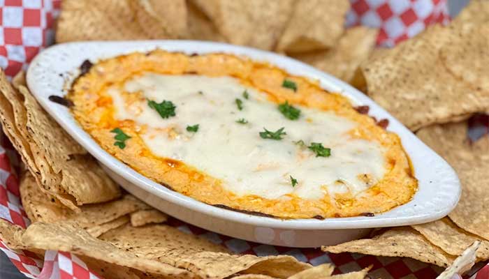 Oval dish of cheesy dip with herbs, surrounded by tortilla chips at a lively restaurant bar.