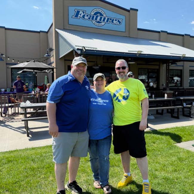 Three people smile on grass in front of a Holmen Wisconsin restaurant with a Features sign.